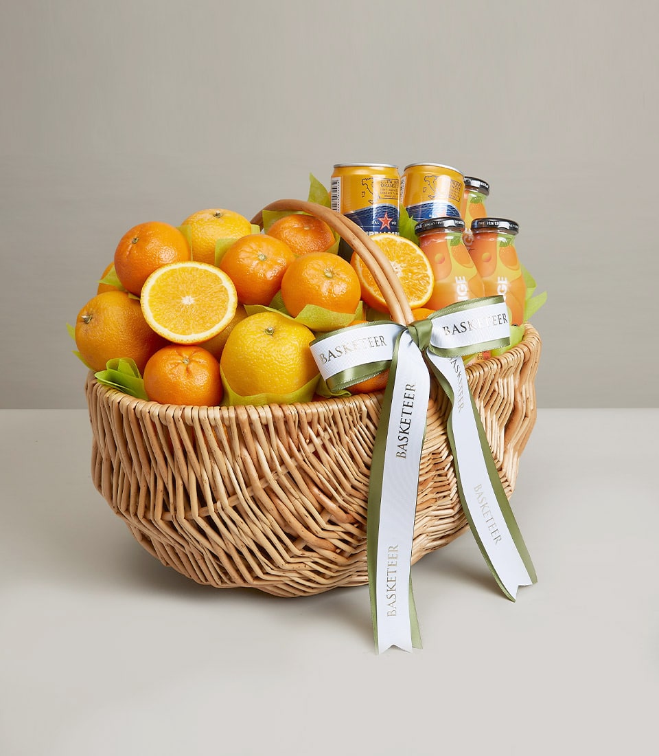Woven wicker basket overflowing with fresh oranges, lemons, and citrus beverages on green tissue, tied with white ribbon