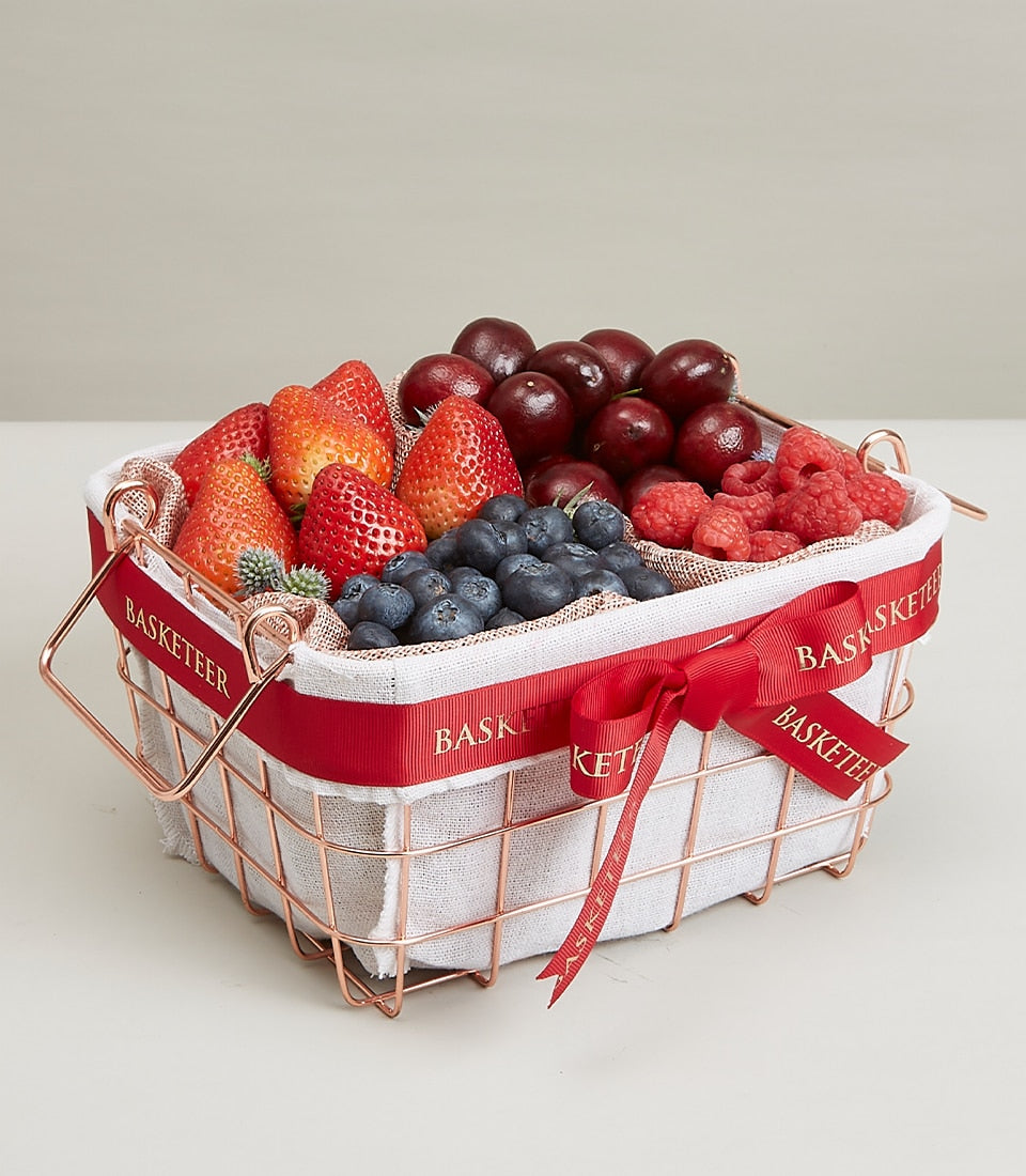 Rose gold wire basket filled with fresh strawberries, blueberries, cherries, and raspberries, tied with red Basketeer ribbon