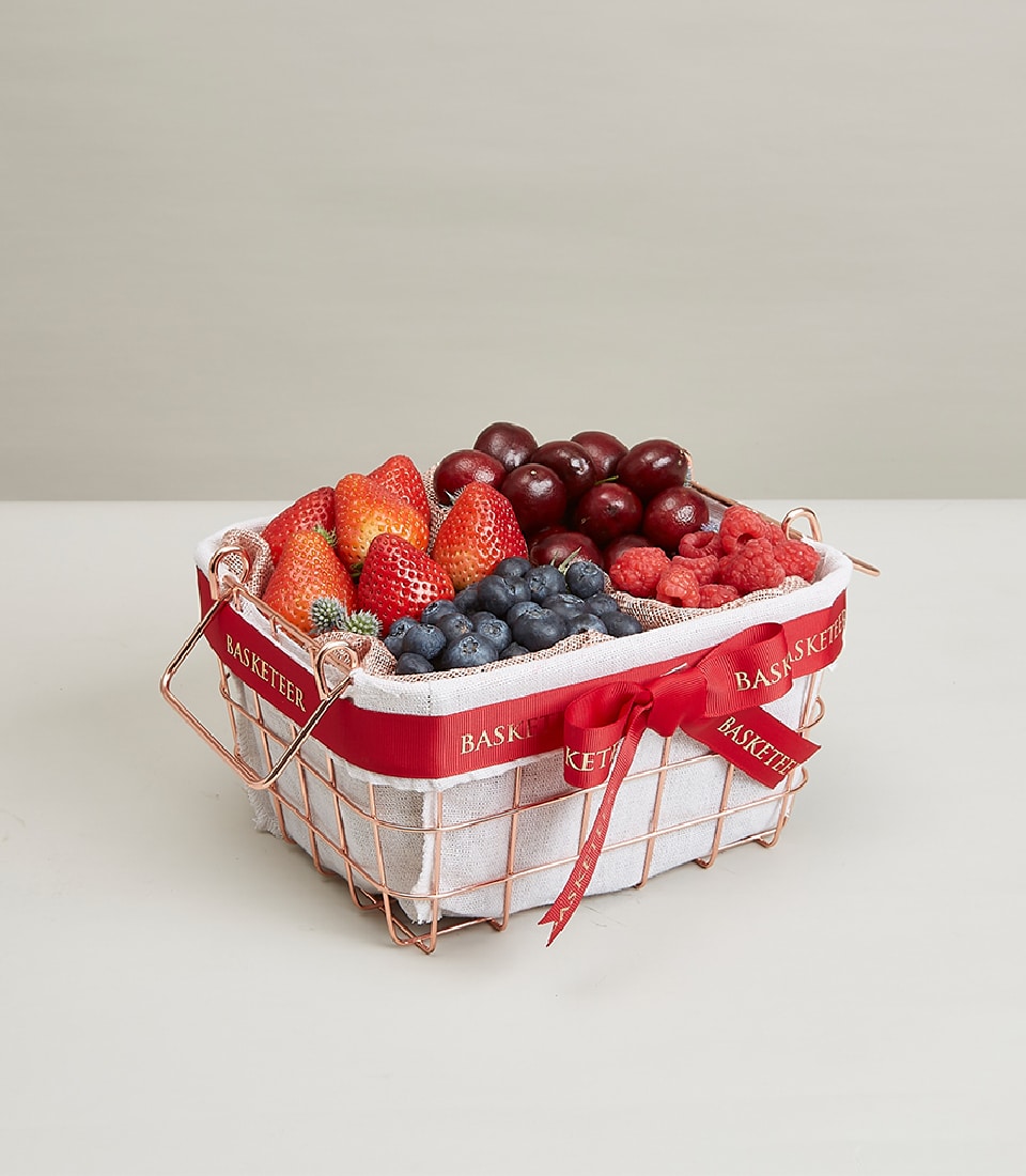 Rose gold wire basket filled with fresh strawberries, cherries, blueberries, and raspberries, tied with red ribbon