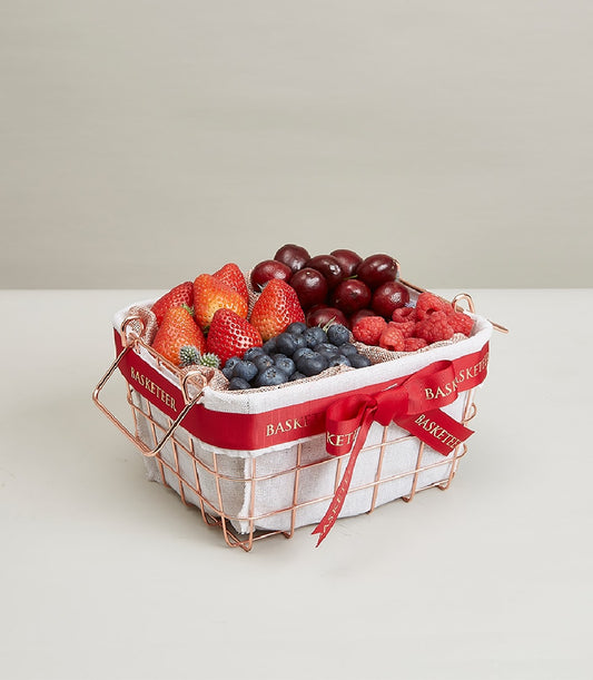Rose gold wire basket filled with fresh strawberries, cherries, blueberries, and raspberries, tied with red ribbon
