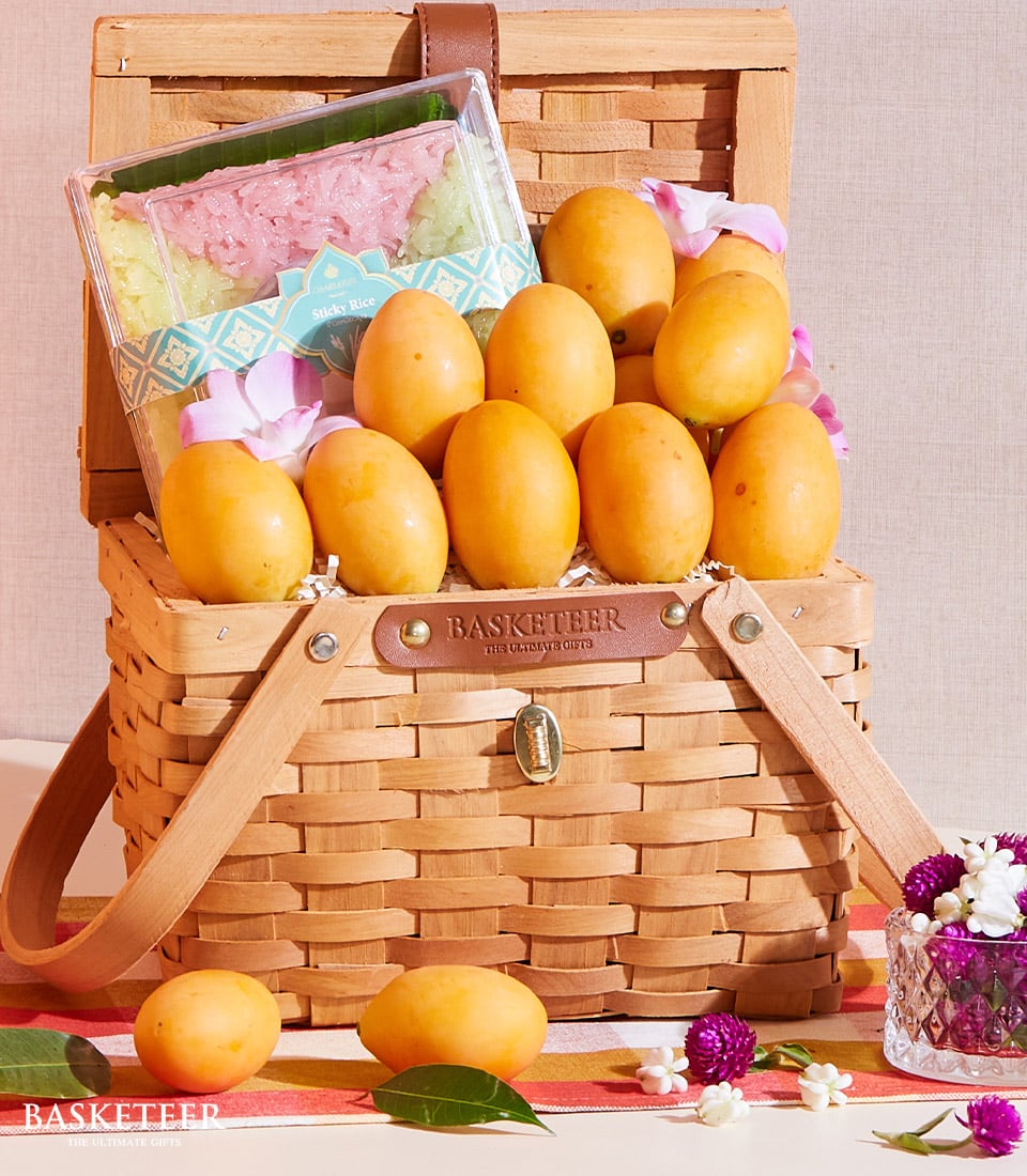 Woven wicker basket filled with fresh yellow plums, sticky rice dessert, and purple flowers for Thai tropical fruit gifting