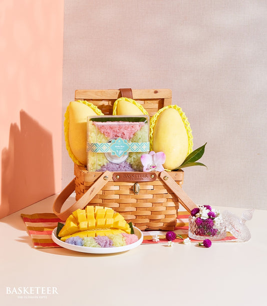 Woven wicker picnic basket with fresh yellow mangoes, layered sticky rice in pink green and purple, and tropical flowers