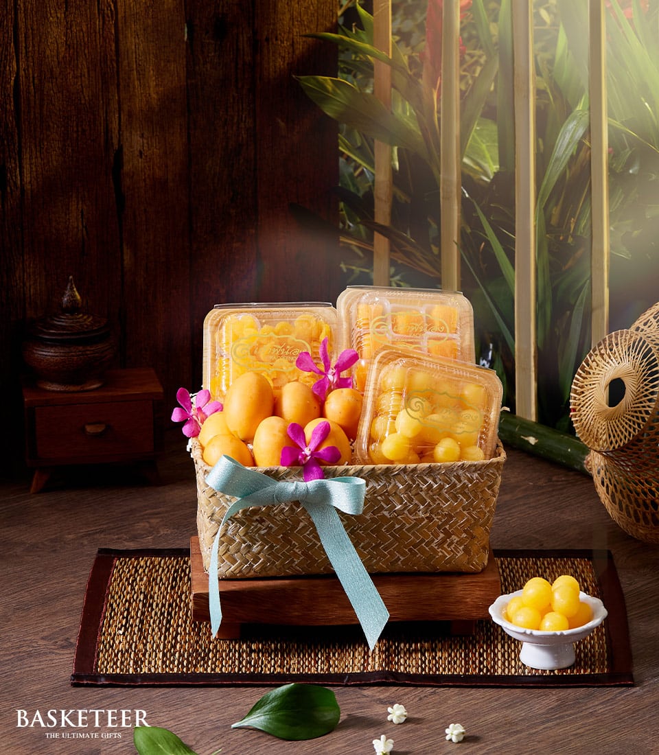 Woven rattan gift basket with Thai desserts, golden sweets, and Marian plums tied with light blue ribbon for Songkran