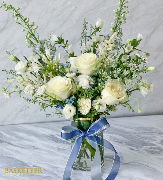Pure white roses and cream blooms in clear glass vase with blue ribbon