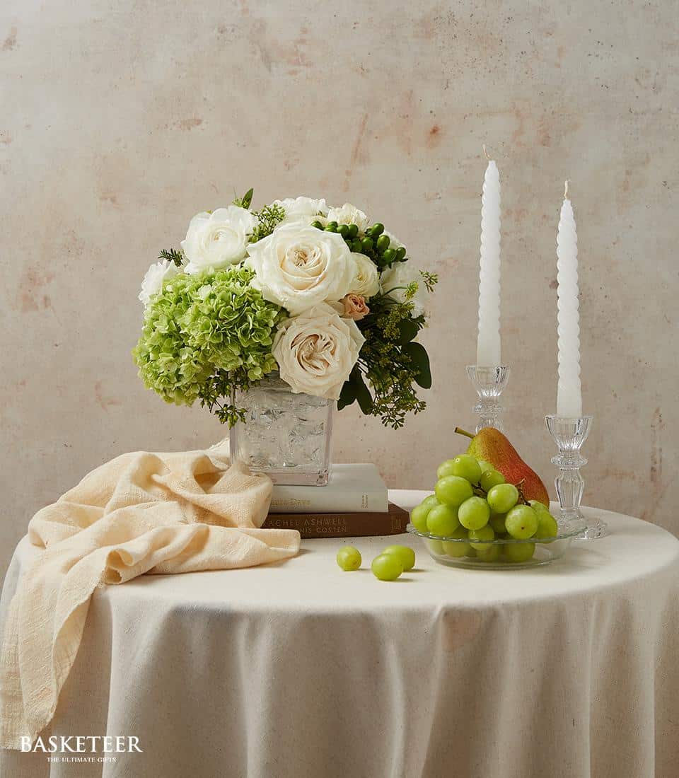 White roses and green hydrangeas in clear glass vase, elegant dining table centerpiece with candles and fruit