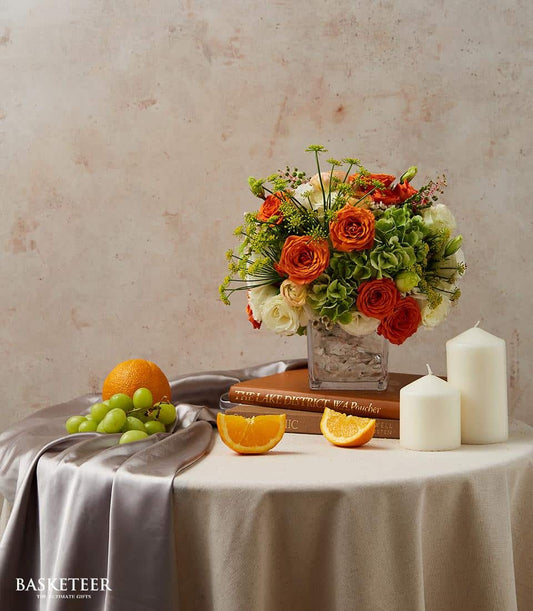 Vibrant orange and white roses with green hydrangeas in a clear glass vase, styled with fresh fruit and candles for dining