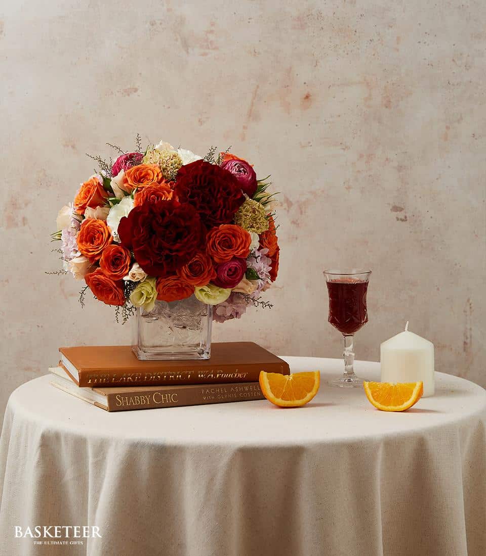Amber Bloom floral arrangement in glass vase with roses and mixed flowers in warm coral and red tones, styled for dining
