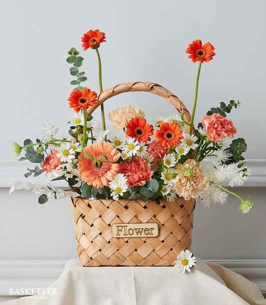 Woven basket of radiant orange gerbera daisies, peach carnations, and white daisies with eucalyptus for get well wishes