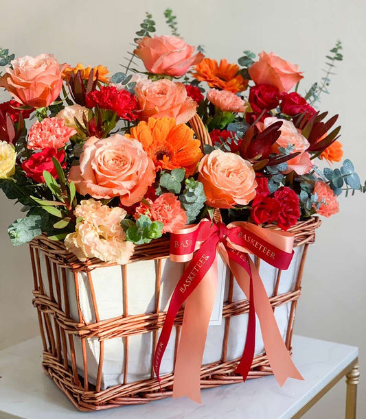 Vibrant mixed roses, gerberas, and carnations in warm orange, peach, and red tones arranged in a wicker basket for get well
