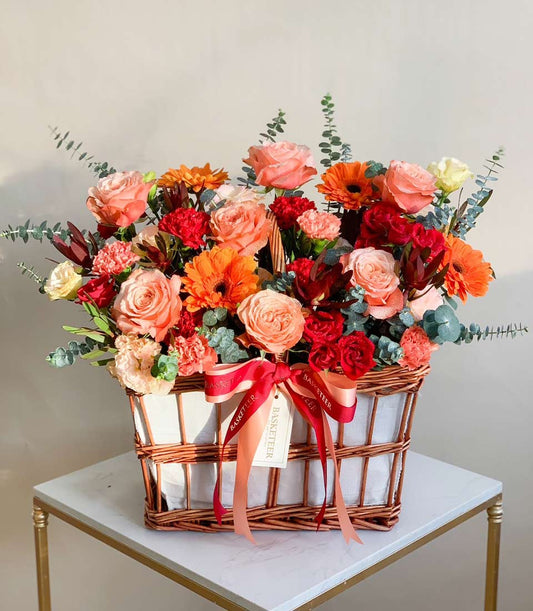 Vibrant peach and coral roses with gerbera daisies in woven wicker basket, get well arrangement with red ribbon bow