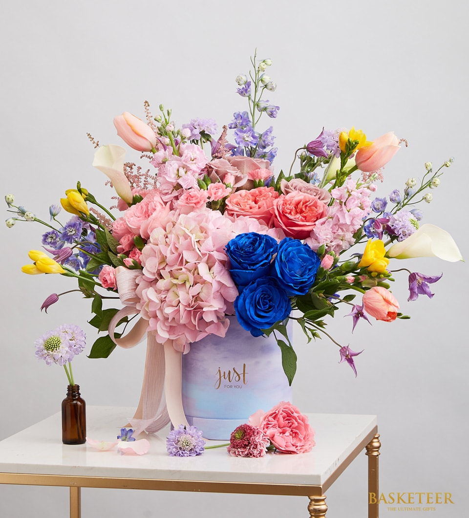 Vibrant mixed flower arrangement in light purple hatbox with pink hydrangeas, blue roses, tulips, and calla lilies