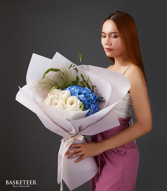 Large bouquet of white and pink roses with vibrant blue hydrangeas, wrapped in white and lavender paper with cream ribbon