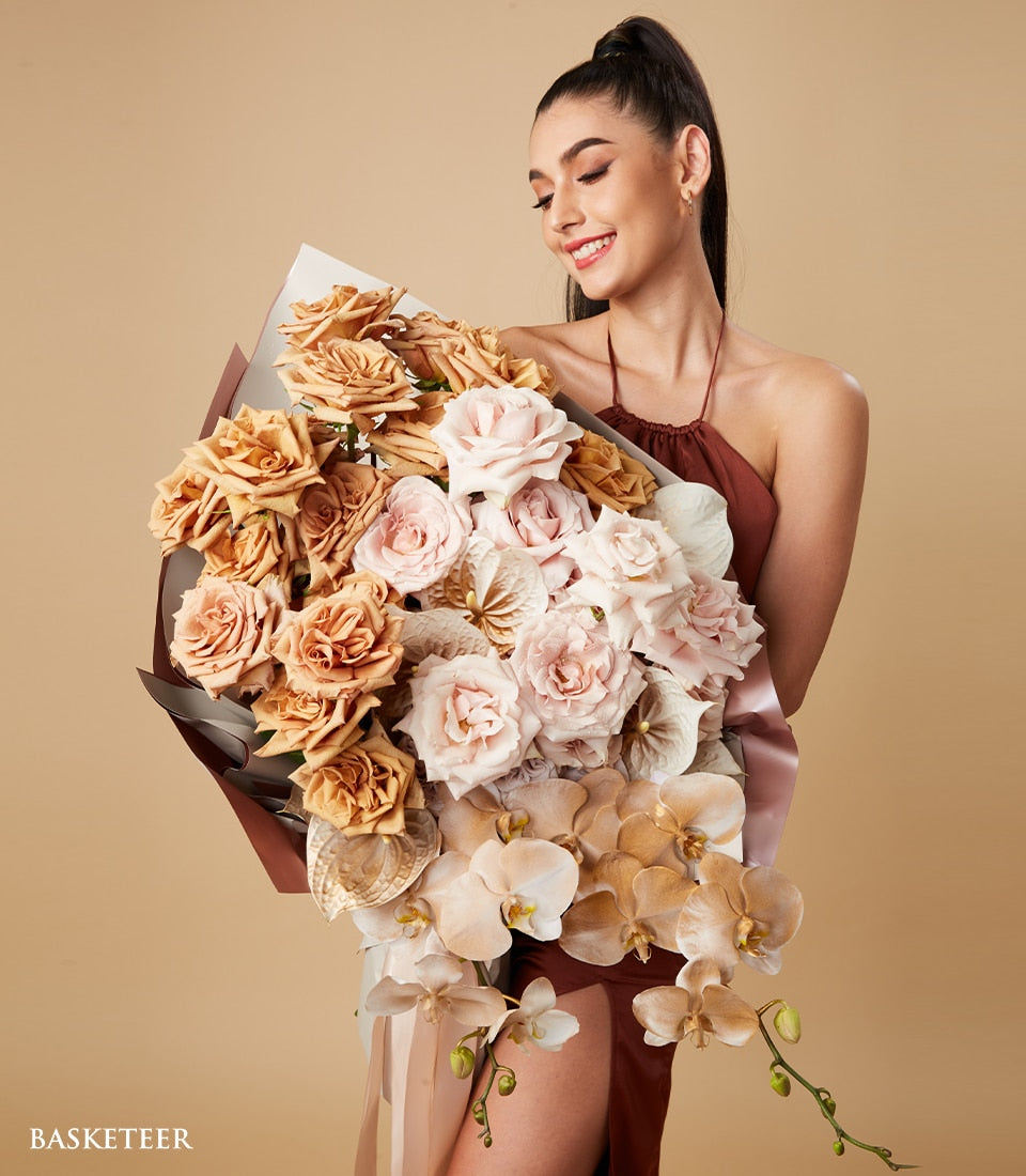 Warm-toned luxury flower bouquet with roses and orchids in cream, blush, and tan, wrapped in brown paper with pink ribbon