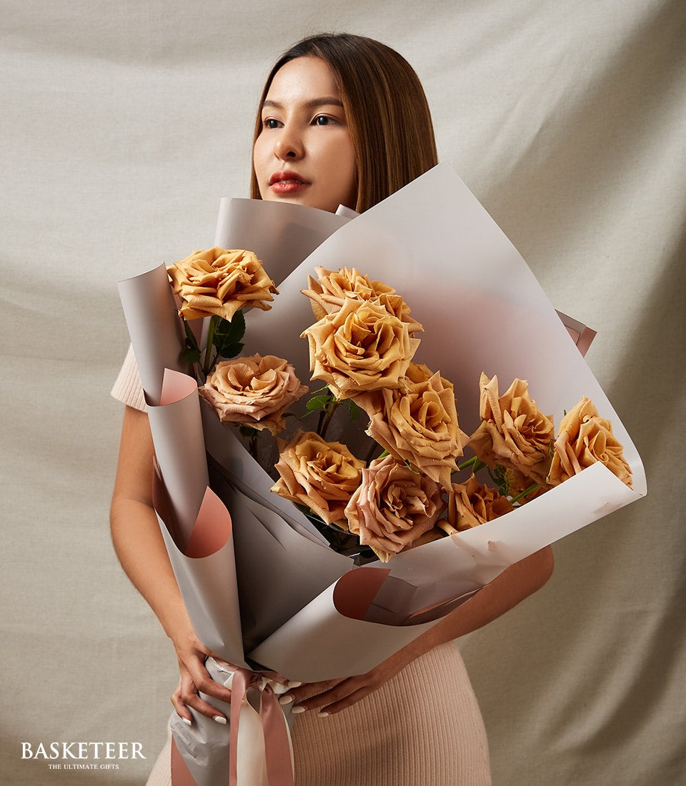 Woman holding premium Toffee Bliss roses bouquet in warm brown and muted orange tones, wrapped in layered grey and peach