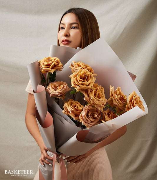 Woman holding premium Toffee Bliss roses bouquet in warm brown and muted orange tones, wrapped in layered grey and peach