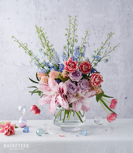 Vibrant mixed flower arrangement with blue delphiniums, pink dahlias, and lavender roses in clear glass vase