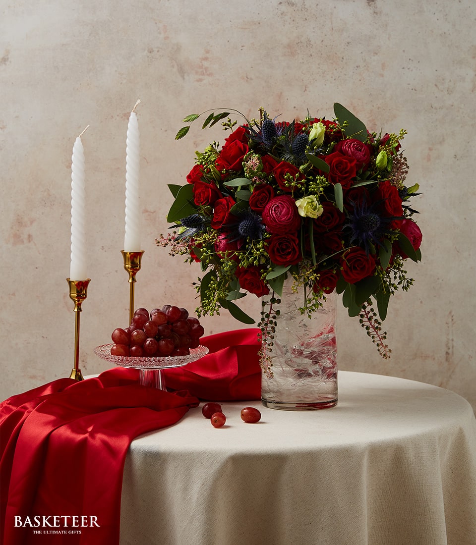 Luxury floral arrangement with red roses, thistles, and greenery in textured glass vase, paired with gold candlesticks and