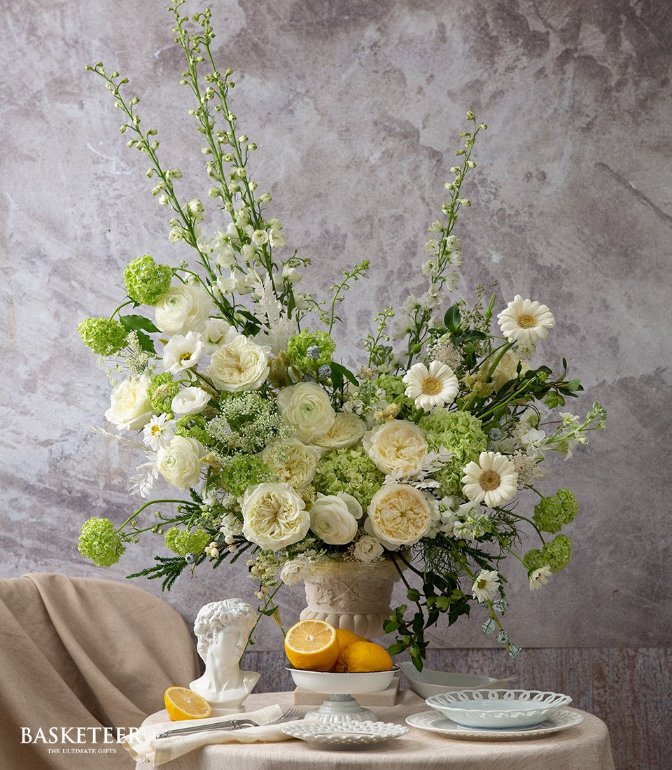 Elegant white and cream rose, ranunculus, and delphinium arrangement in ornate ceramic vase with green foliage