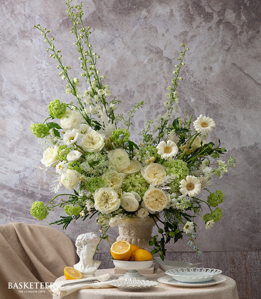 Elegant white and cream rose, ranunculus, and delphinium arrangement in ornate ceramic vase with green foliage