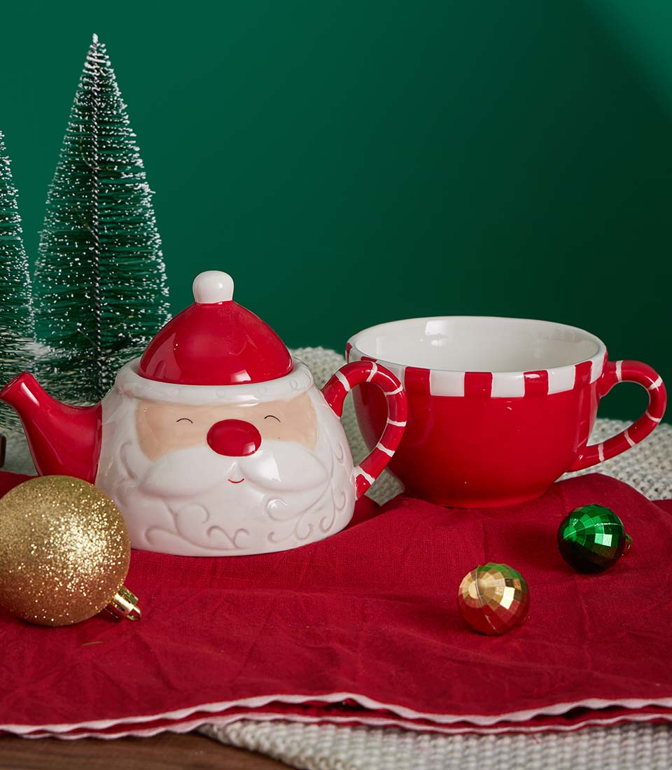 Festive Christmas tea set with Santa teapot, striped mug, gourmet cookies and chocolates on red napkin with gold ornaments