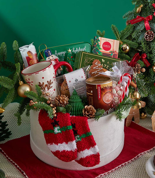 Festive Christmas gift basket with hot chocolate, cookies, reindeer mug, and holiday treats on dark green backdrop