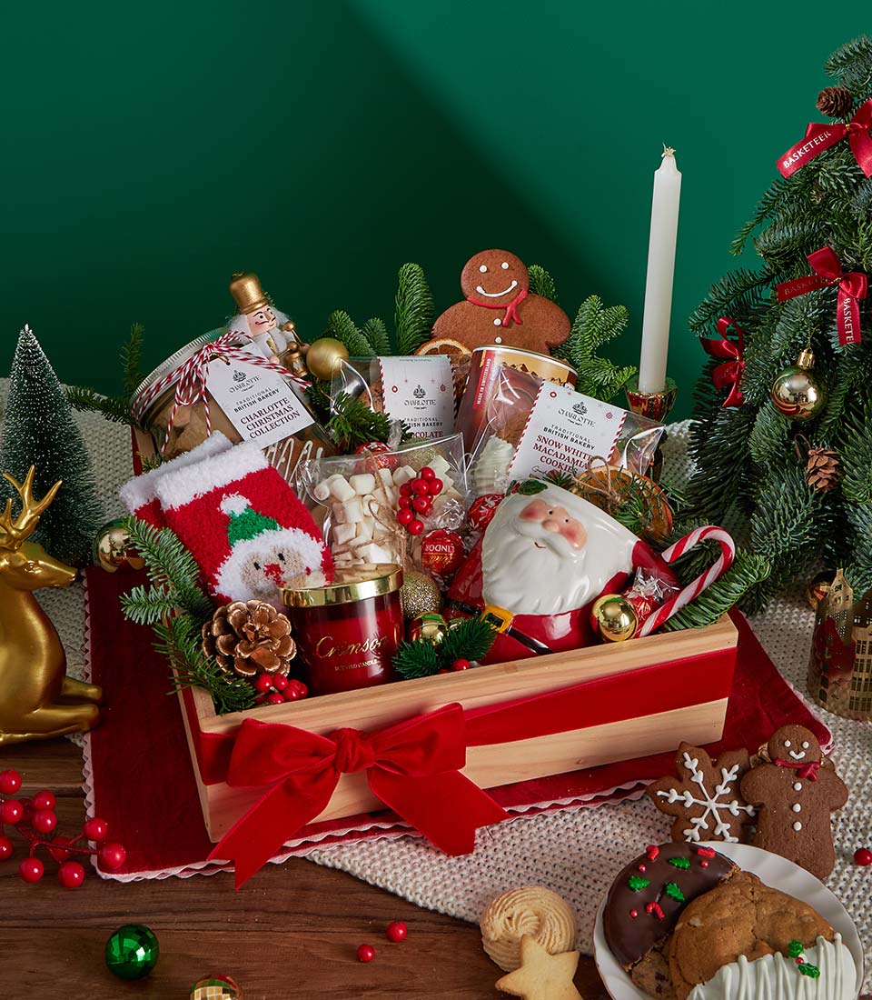 Festive Christmas cookie and hot chocolate gift basket in wooden crate with red velvet bow, Santa mug, and holiday treats