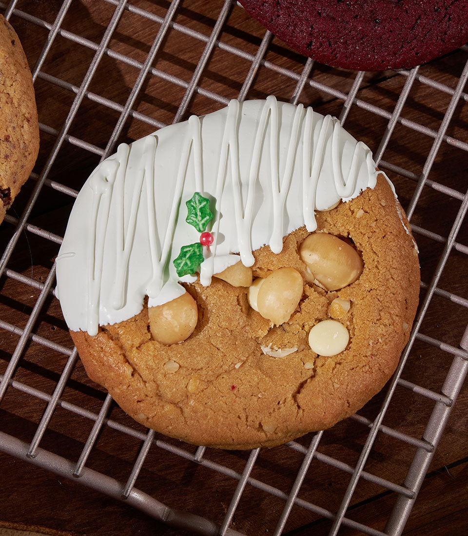 Soft-baked Christmas cookies with macadamia nuts, white chocolate icing, and festive holly garnish on cooling rack