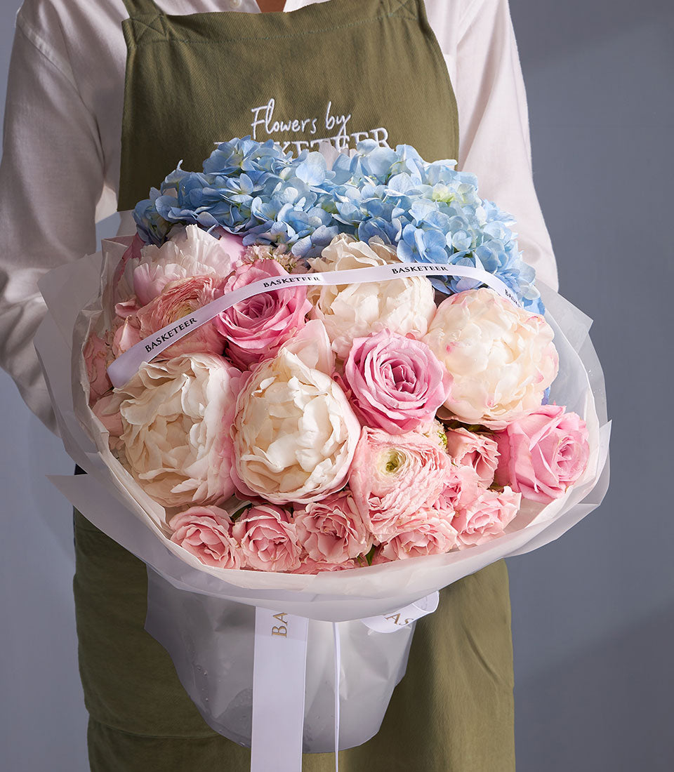 Pastel bouquet with light blue hydrangeas, cream peonies, and pink roses wrapped in white paper