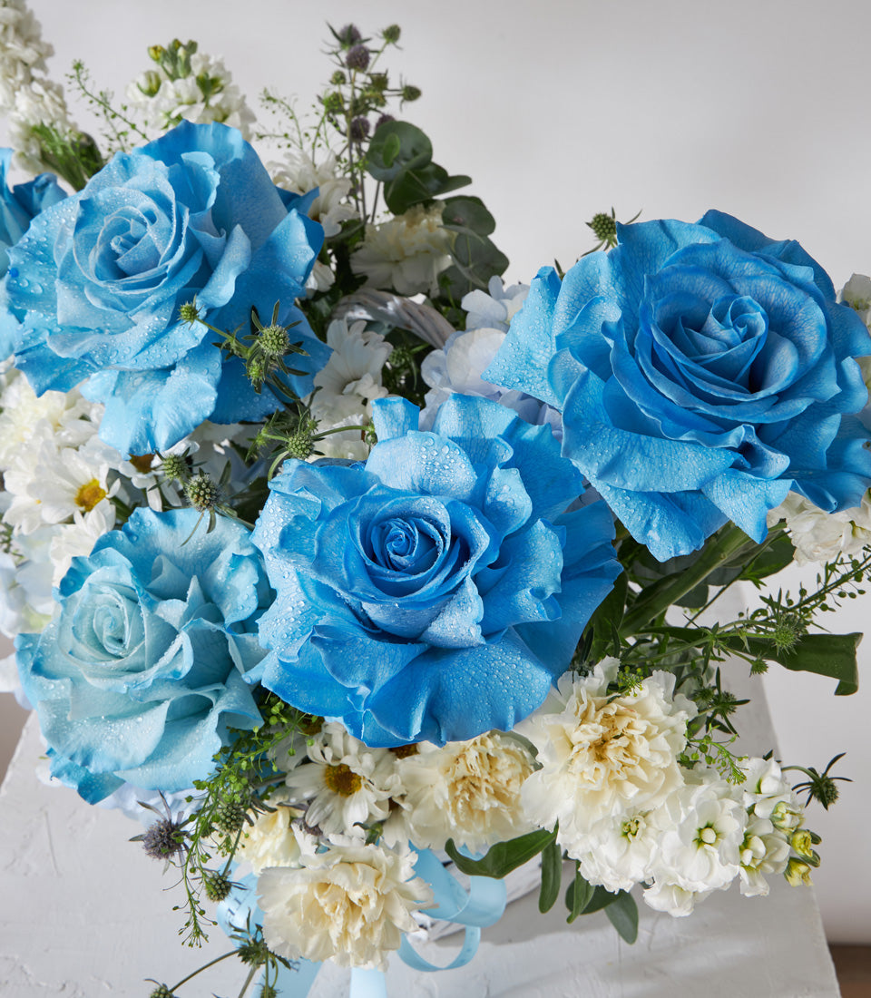 Blue and white rose bouquet with daisies and carnations in white woven basket with light blue ribbon