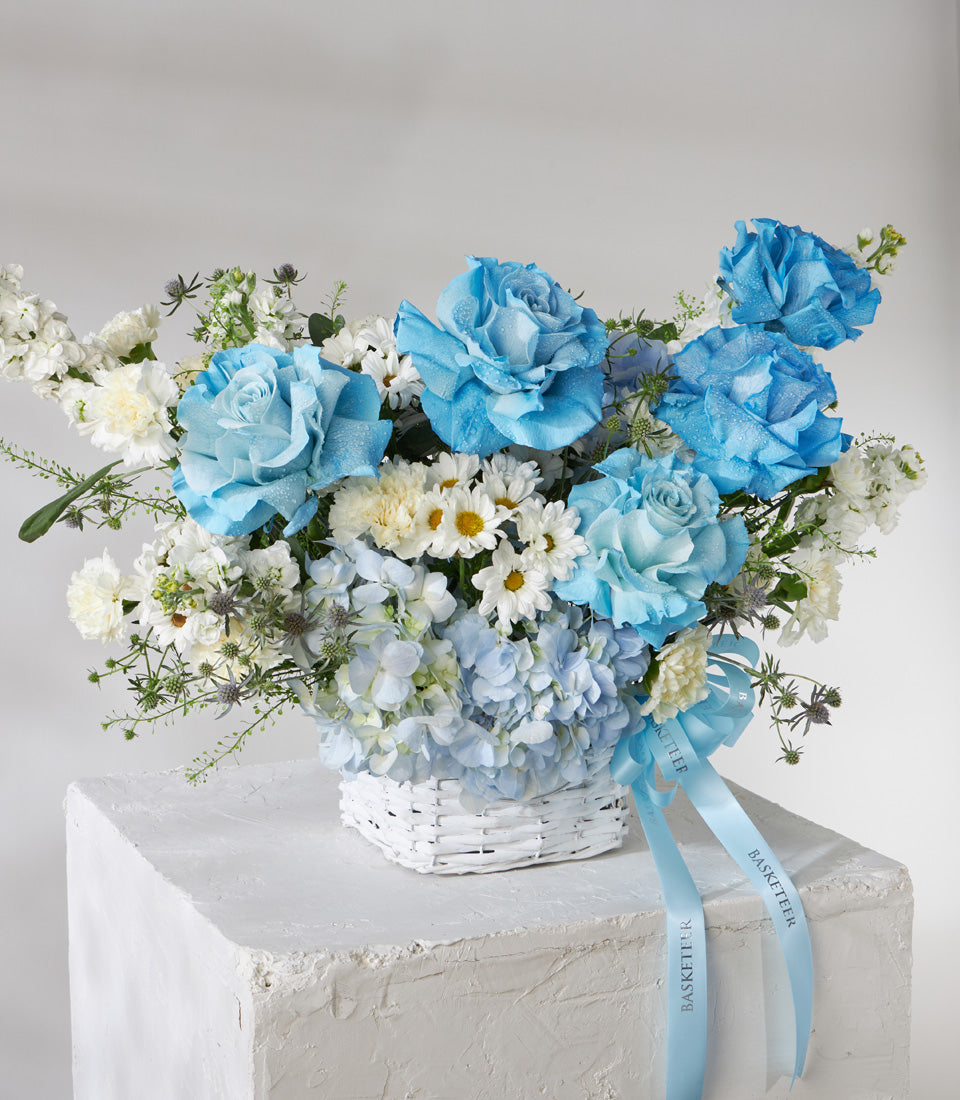 Blue roses and white flowers in white woven basket with fresh arrangement of hydrangeas and daisies