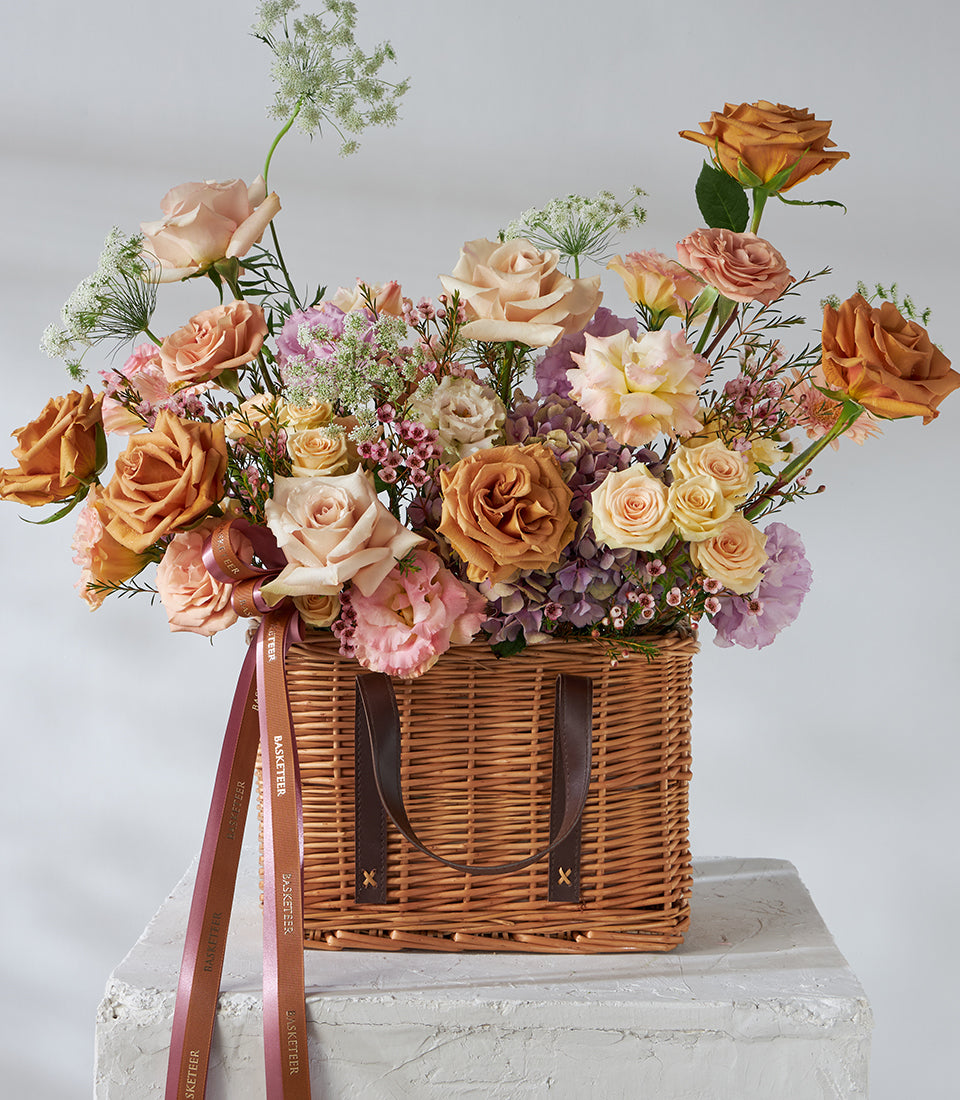 Caramel and blush roses with lilac flowers in woven wicker basket, tied with branded ribbons