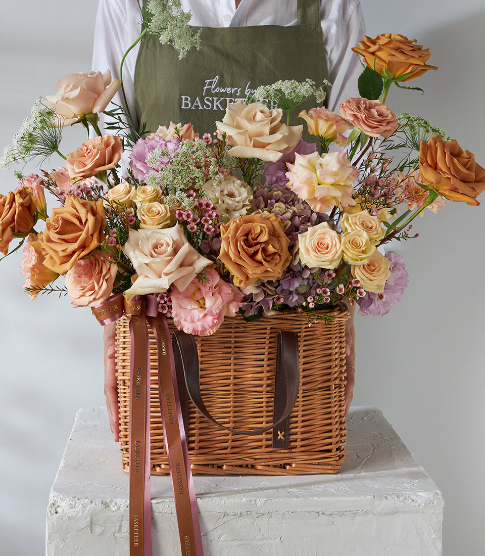 Caramel and blush rose arrangement in lilac wicker basket with hydrangeas and ribbon
