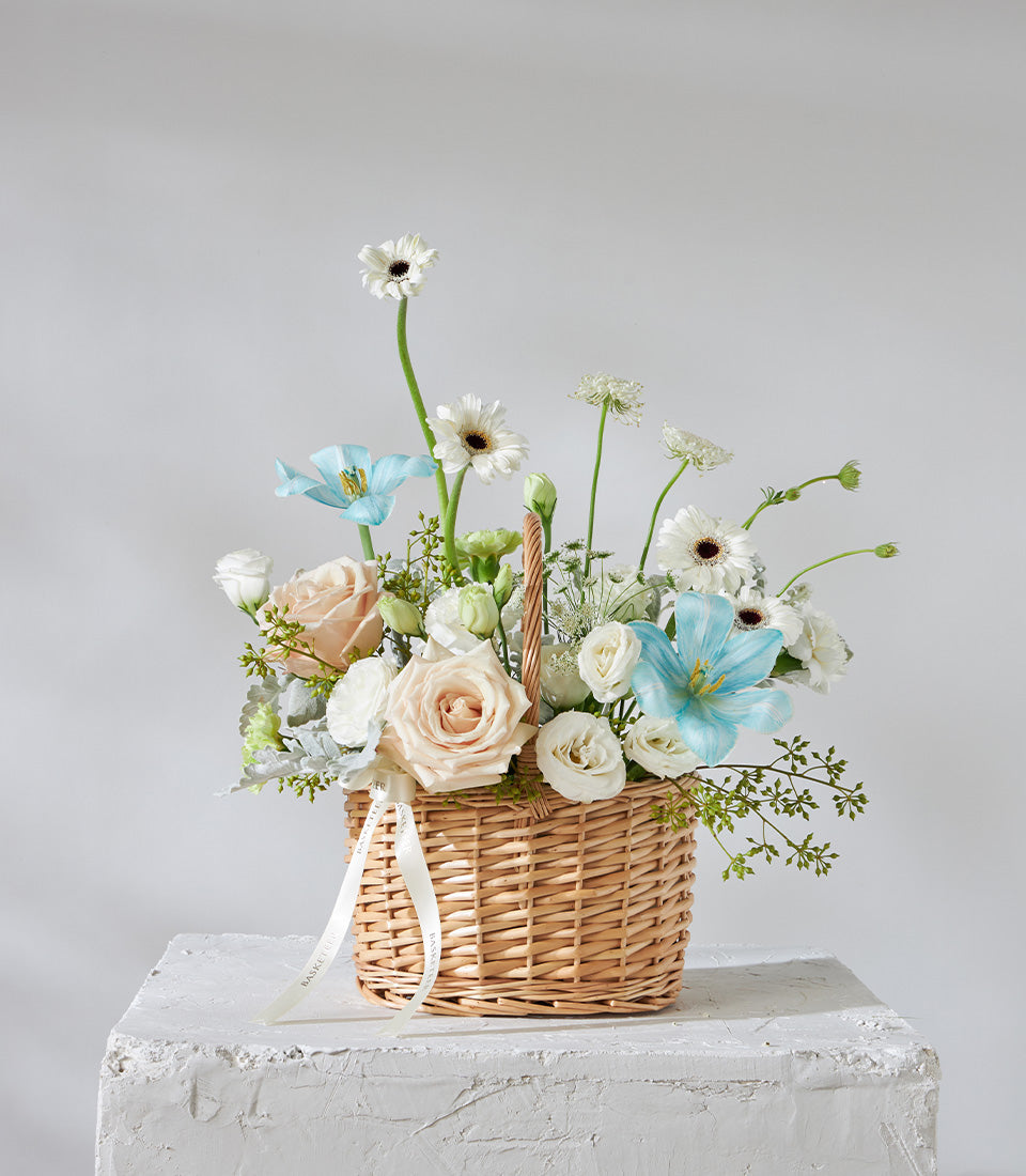 Light blue and blush woven basket with white roses, peach roses, and blue tulips, tied with cream ribbon for birthday