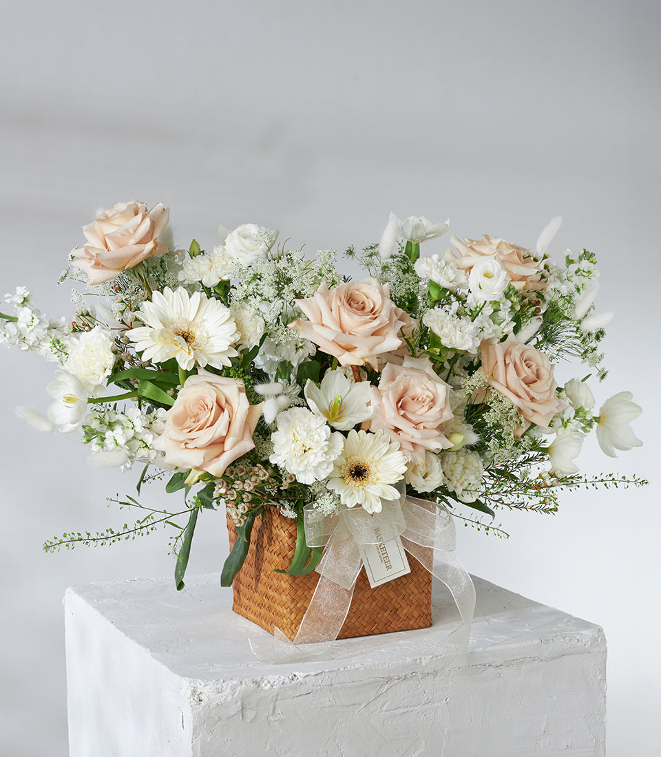 Cream roses and white gerberas in woven basket with trailing greenery and beige ribbon bow
