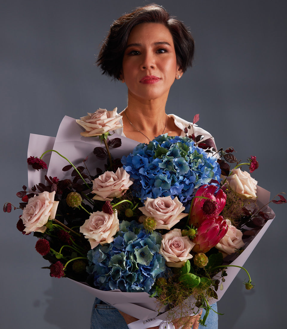 Luxury bouquet of blue hydrangeas, pink roses, and red protea with dark green foliage in purple wrapping