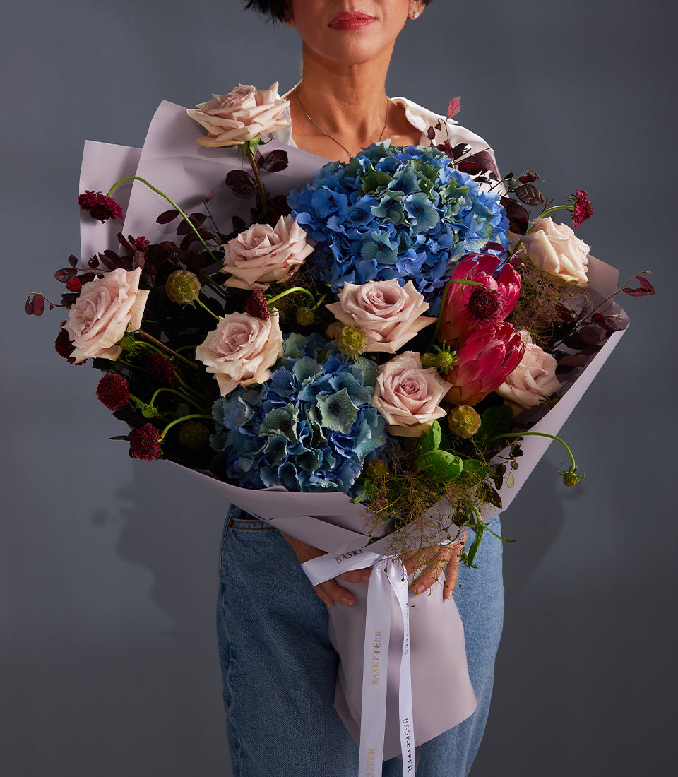 Elegant luxury bouquet with light pink roses, blue hydrangeas, and fuchsia protea, wrapped in lavender paper