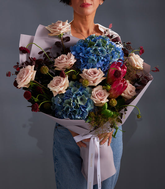 Elegant luxury bouquet with light pink roses, blue hydrangeas, and fuchsia protea, wrapped in lavender paper
