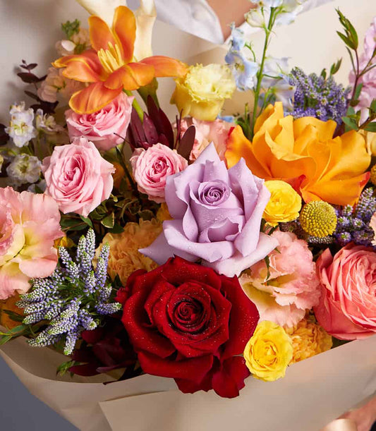 Vibrant mixed bouquet with orange lilies, pink and red roses, lavender blooms and yellow accents in kraft paper wrapping