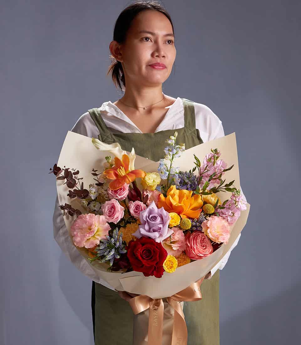 Woman holding vibrant mixed bouquet with orange lilies, roses, and pastel wildflowers in cream paper wrap