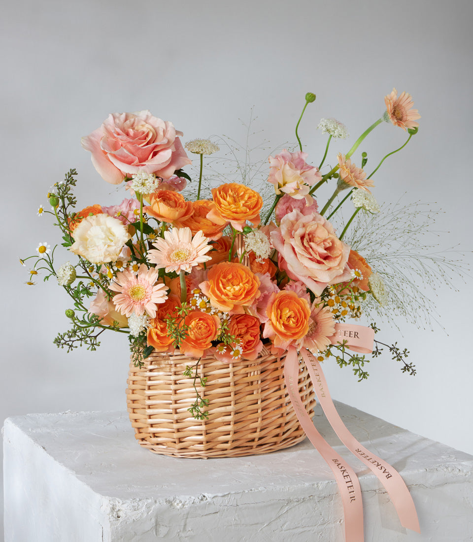 Sunset Glow peach and orange rose basket with gerbera daisies, cream blooms, and wispy greenery in woven wicker