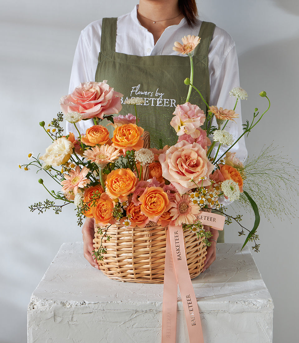 Florist holding round wicker basket with peach roses, pink garden roses, and orange gerberas tied with Basketeer ribbons
