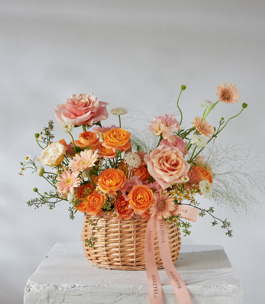 Sunset Glow premium flower basket with roses, ranunculus, and gerbera daisies in warm orange and peach tones, tied with pink