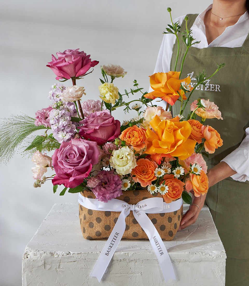 Sunset orange and mauve rose basket with ranunculus and daisies in burlap carrier, ready for same-day Bangkok delivery
