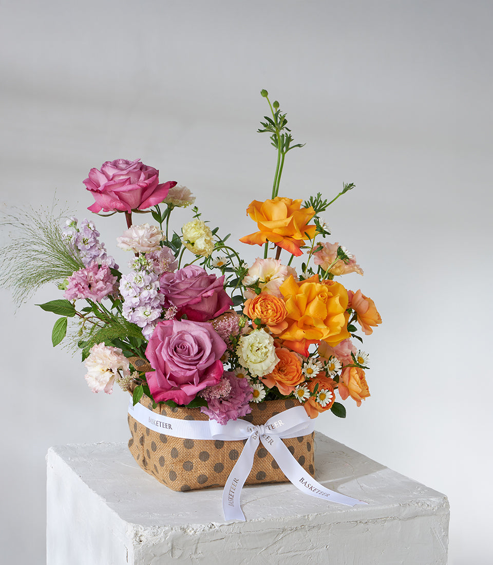 Sunset orange and mauve roses in rustic polka-dot basket with mixed blooms and white ribbon