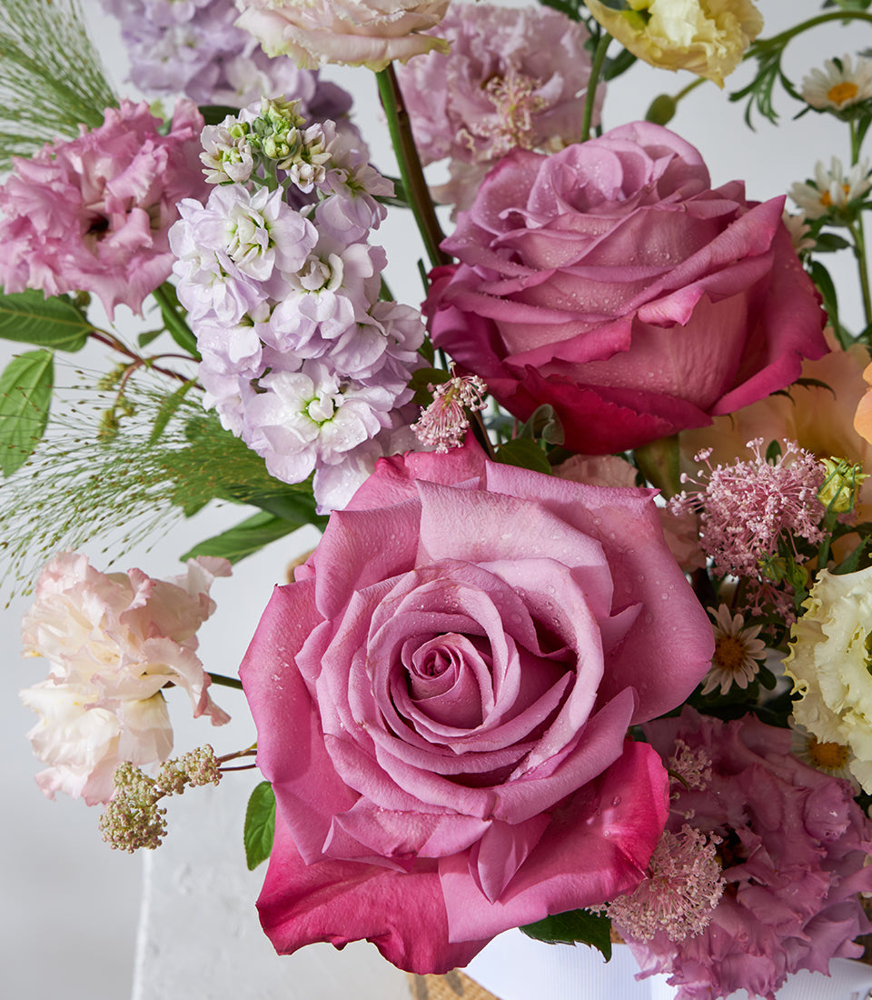 Sunset orange and mauve rose basket with stock flowers, carnations, and lisianthus in white woven container