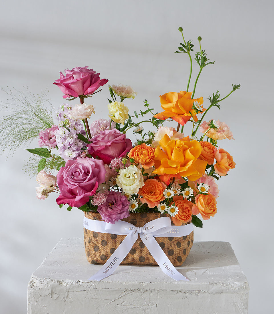 Sunset orange and mauve roses in rustic basket with white ribbon, mixed with purple and yellow blooms