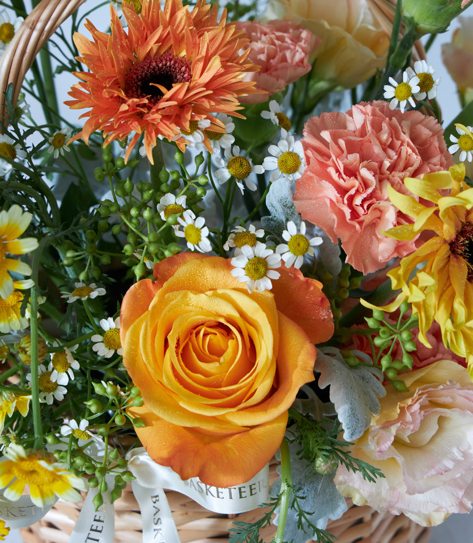 Vibrant birthday flower basket with orange roses, gerberas, pink carnations, and sunflowers in woven wicker