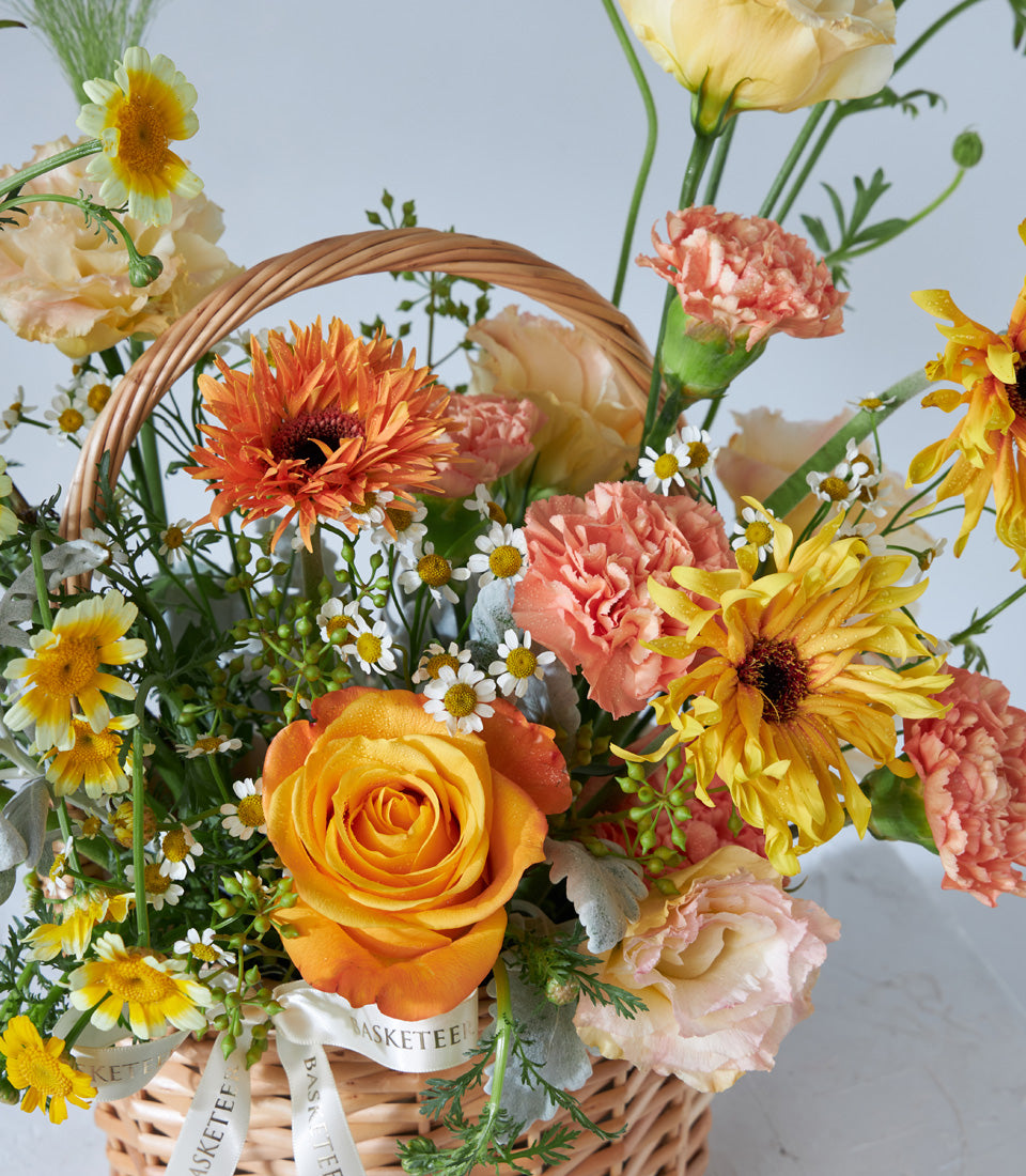 Vibrant golden gerbera, sunflower, and rose arrangement in woven wicker basket with white ribbon, birthday flower gift
