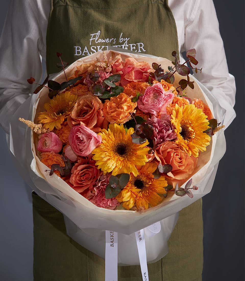 Florist holding vibrant mixed bouquet with yellow gerberas, orange roses, pink ranunculus, and deep red carnations wrapped
