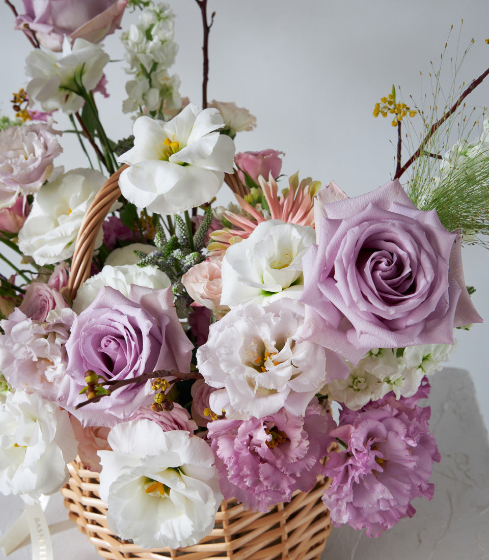 Lavender Grace wicker basket with soft purple roses, white lisianthus, and pink blooms